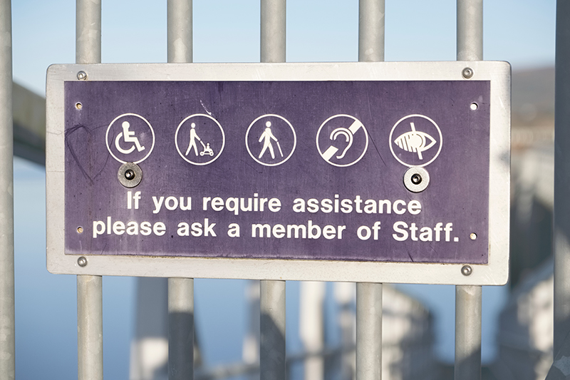 Sign offering assistance to people with disabilities with a wheelchair, stroller, cane and hearing and visual impairment logos.