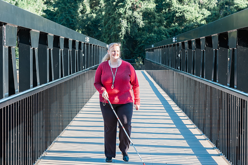 Blind female walks across a bridge using a long white cane.